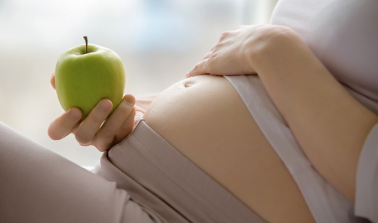 Беременная женщина одной рукой держится за живот, а в другой руке держит яблоко